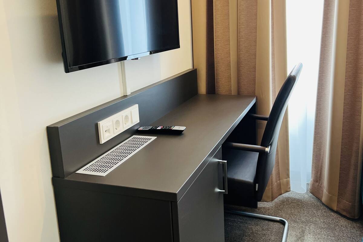 Habitación de hotel con un televisor de pantalla plana montado en un escritorio de color oscuro, un enchufe y un control remoto. Se observa una silla con estructura metálica y piso alfombrado.