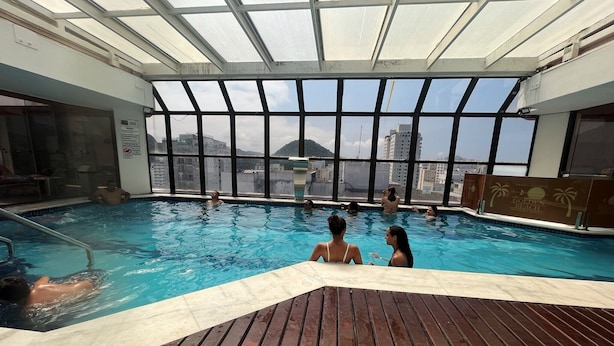 An indoor swimming pool with a view of the cityscape through large windows. - Apartment with beautiful sea view - Praia de Pitangueiras (Guarujá)