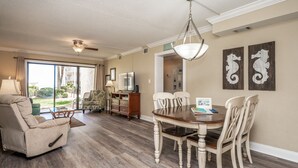 A spacious living room with a dining area, a ceiling fan, and a large window providing a view of the outdoors. - Castle Reef 123 beautiful beachfront condo! (New Smyrna Beach)