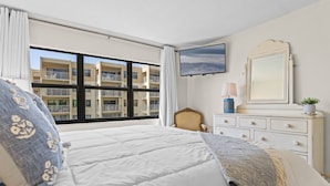 Ein Schlafzimmer mit einem großen Bett, einem Schminktisch, einem Sessel, einem Fernseher und einem Fenster mit Blick auf Gebäude.