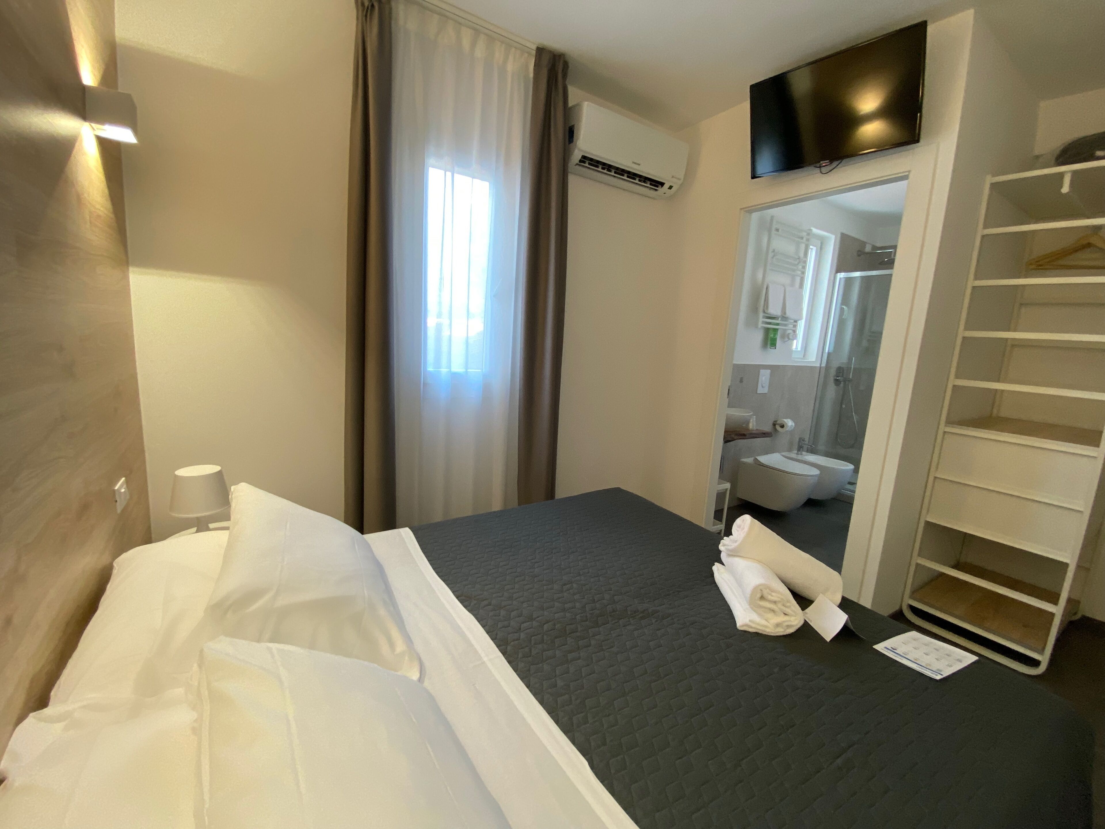 Een hotelkamer met een bed, een televisie, een airconditioning, een kast en een badkamer, zichtbaar in een spiegel.