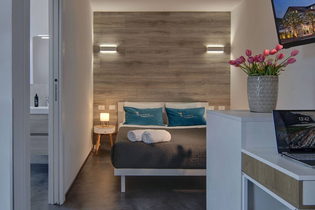 Una habitación de hotel moderna con una cama, una mesita de noche, una computadora portátil y un florero con flores.
