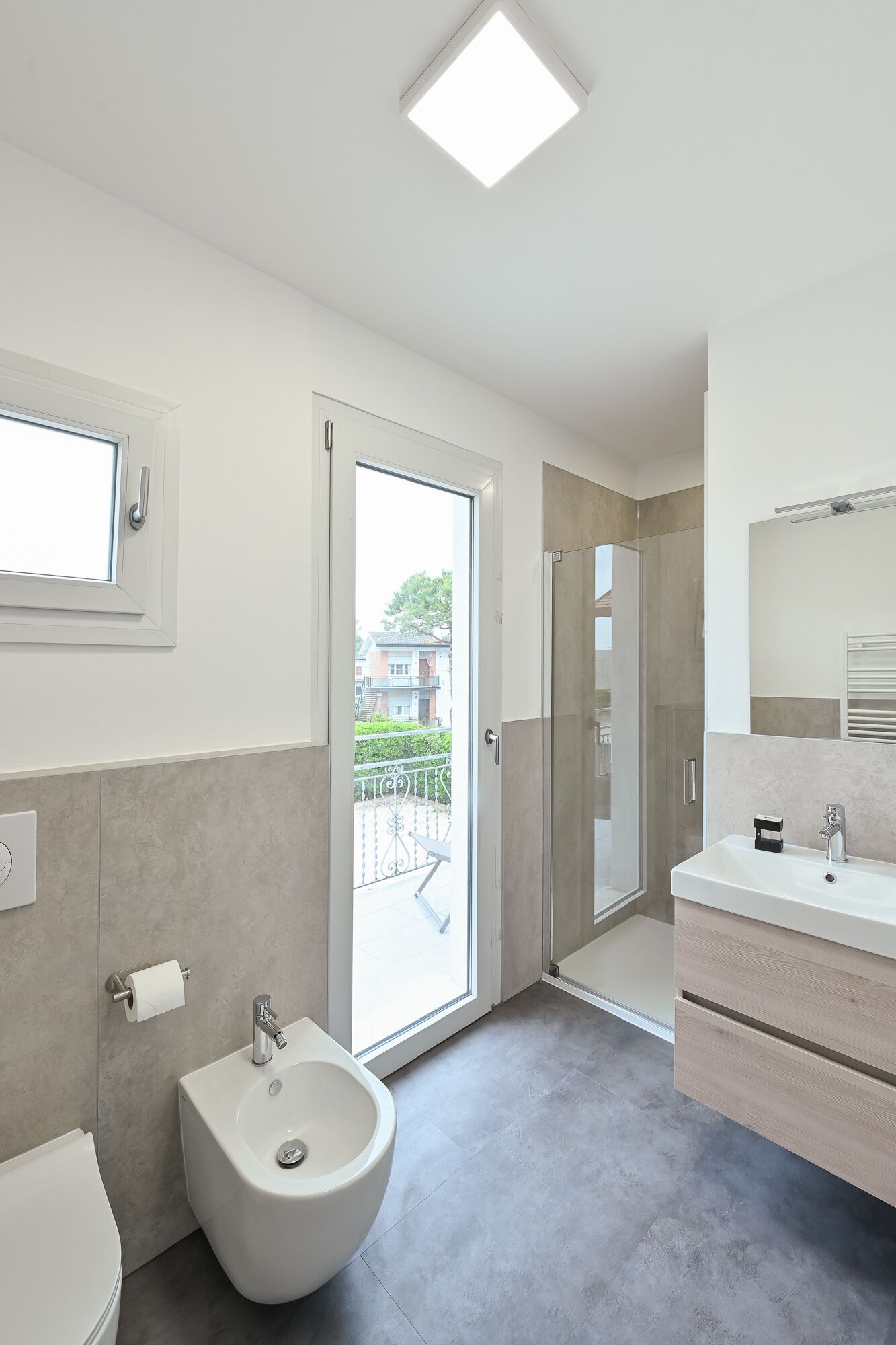 A modern bathroom with a bidet, toilet, and sink. There is a large window and a door leading to an outdoor balcony.