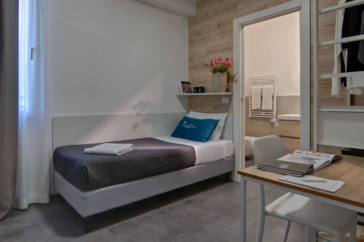Una cama individual con un almoháron azul, una mesita de noche con un cuaderno, bolígrafo y una computadora portátil.