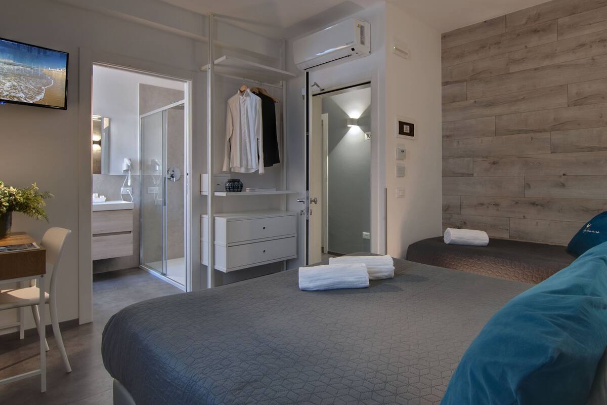 Habitación de hotel con cama, escritorio, silla, baño con ducha de vidrio y televisor montado en la pared.