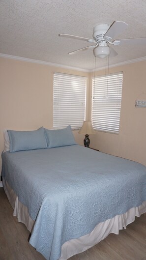 A bedroom with a bed, two windows with blinds, a ceiling fan, and a bedside lamp.