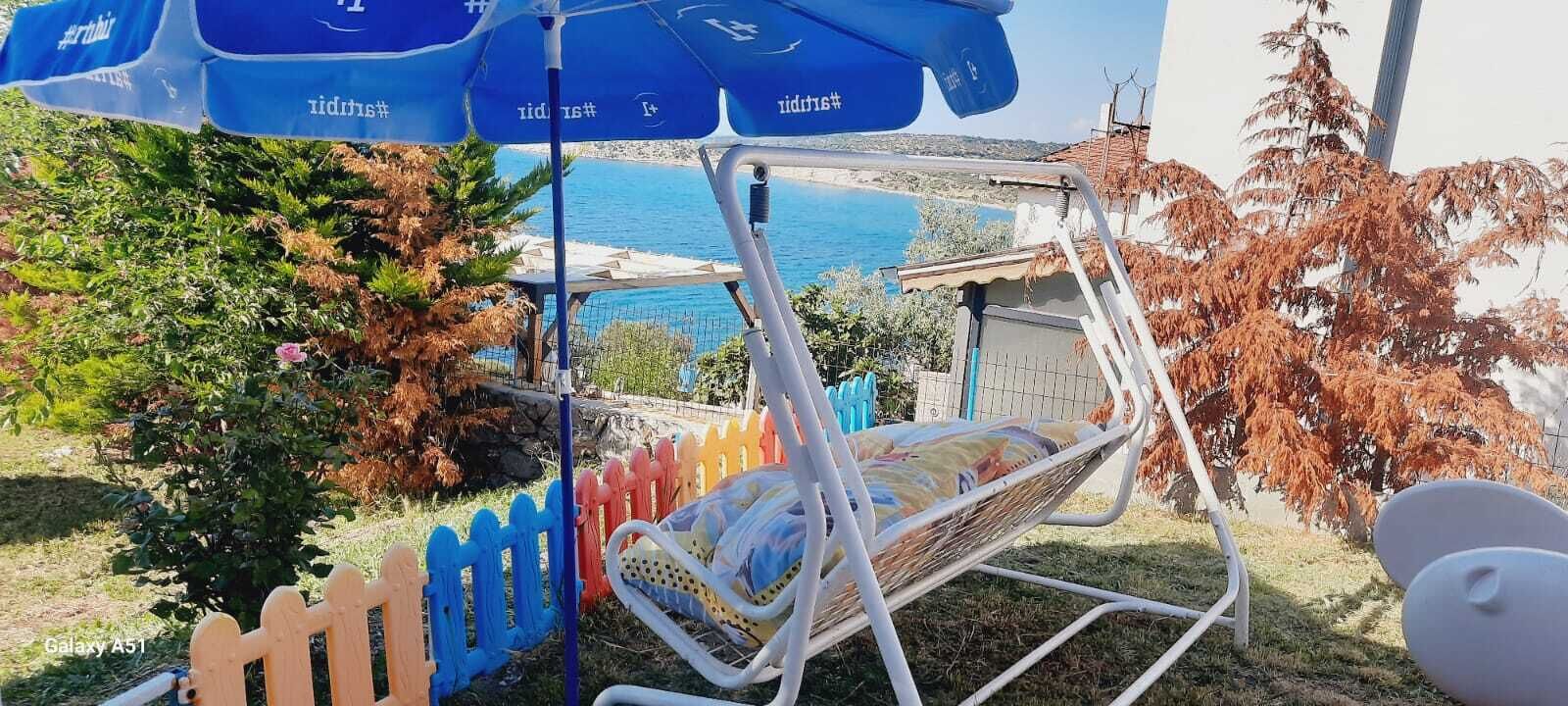 Uma cadeira rede com vista para a área costeira, uma cerca de madeira e um guarda-sol azul com texto.