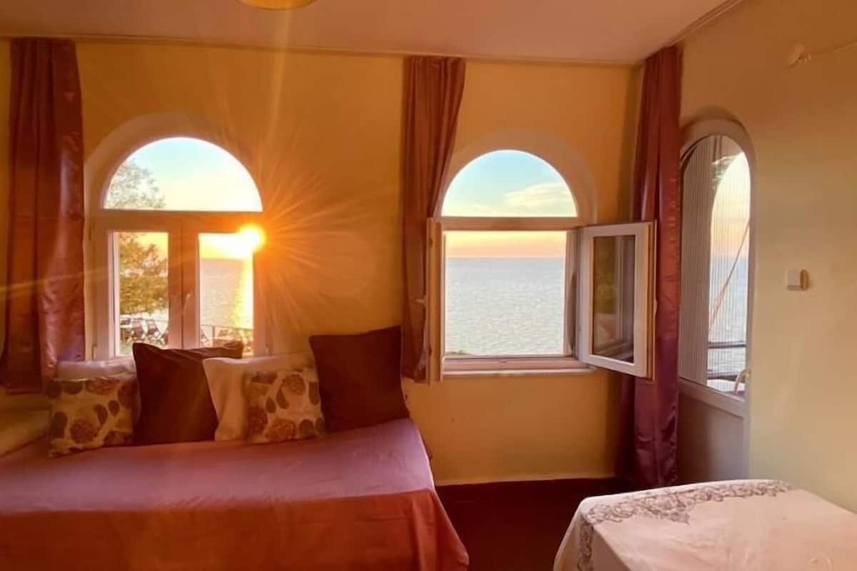 A room with a bed, a desk, and two windows with views of the sea.