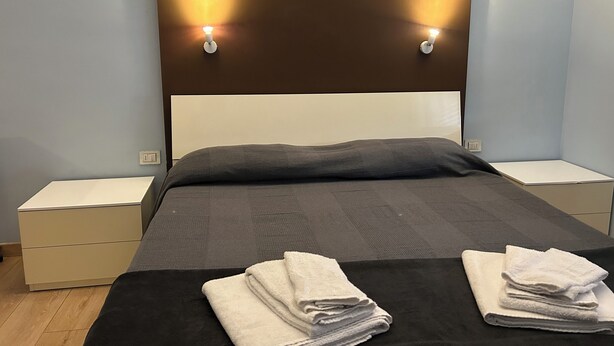 A neatly made bed with a dark headboard, two bedside tables, and a wall-mounted light fixture. - Ancora Residence - Downtown Area - Hospital (Piacenza)