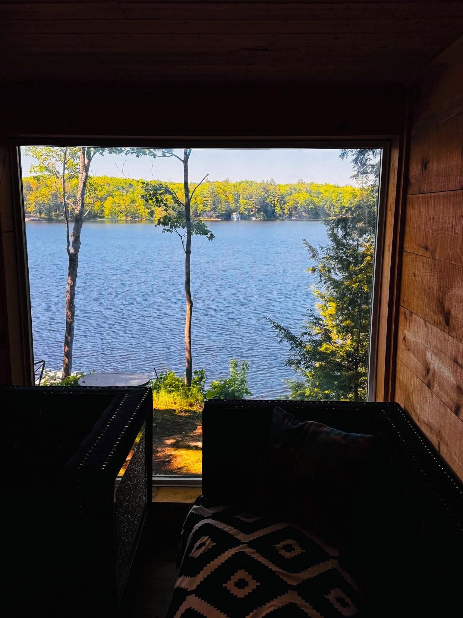 Ein Zimmer mit Blick auf einen See und Bäume, mit Holzboden und einem gemusterten Kissen.