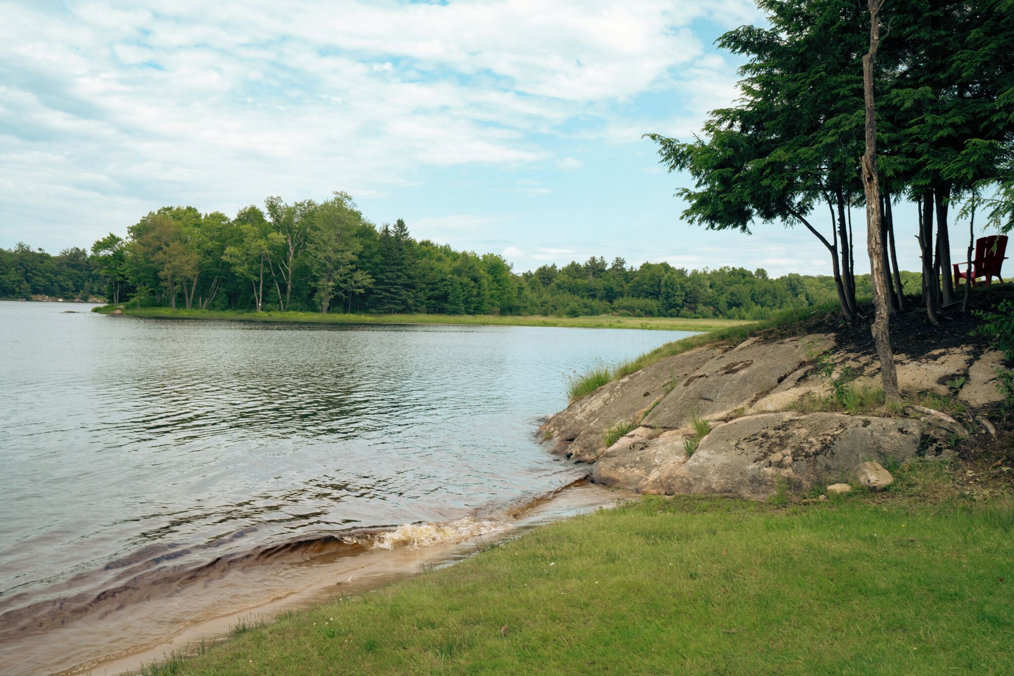 A lakeside view with a grassy shore, trees, and a red chair.