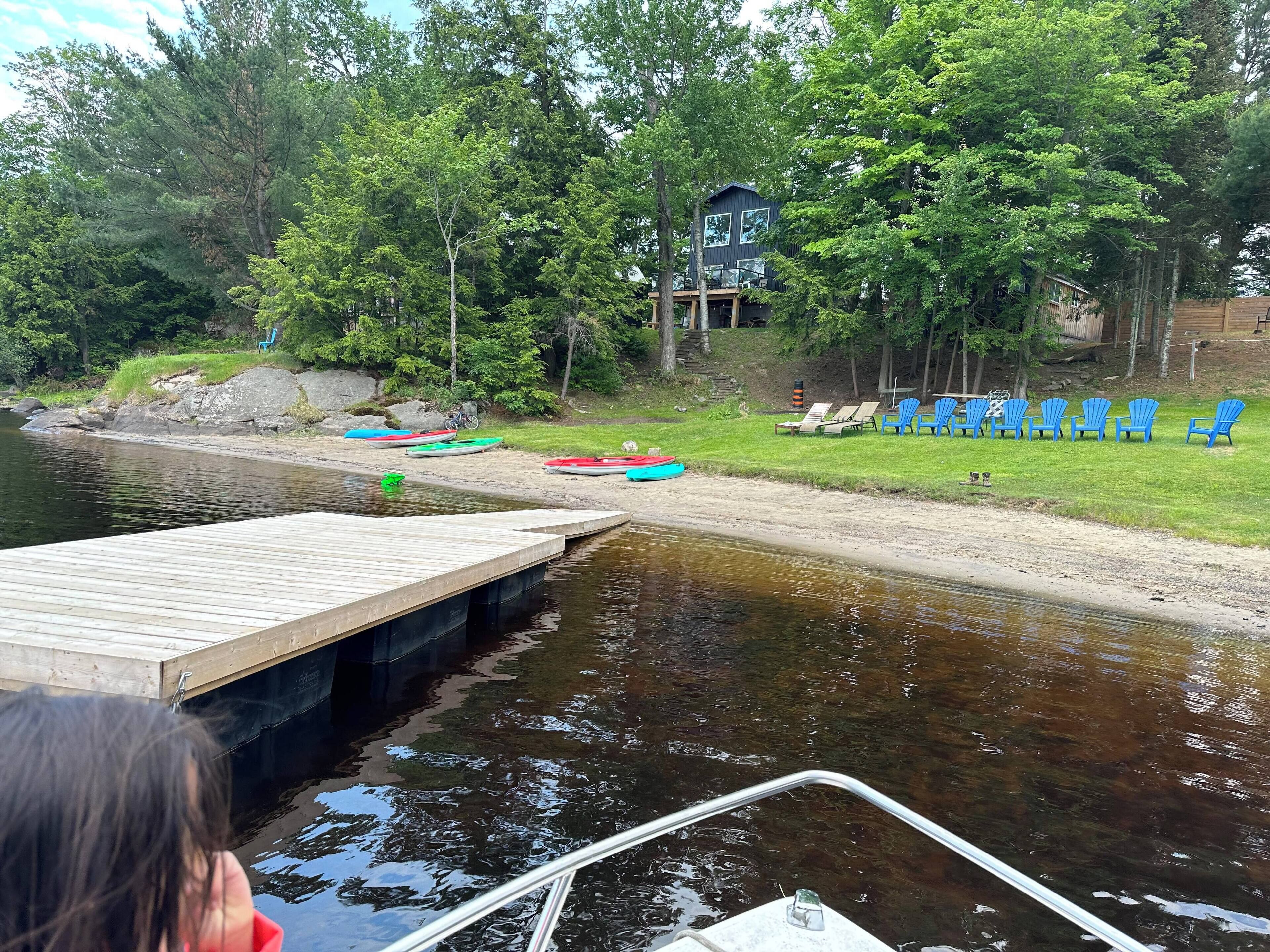 A wooden dock extending into a calm lake, surrounded by lush greenery and a small building with a balcony.