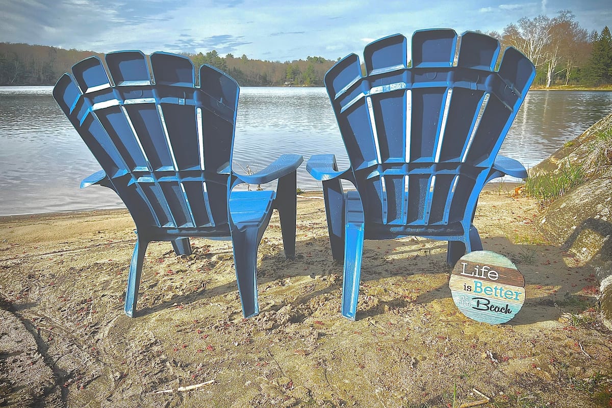 Deux chaises Adirondack bleues sont placées face à un lac, et un panneau indique : « La vie est plus belle à la plage. »