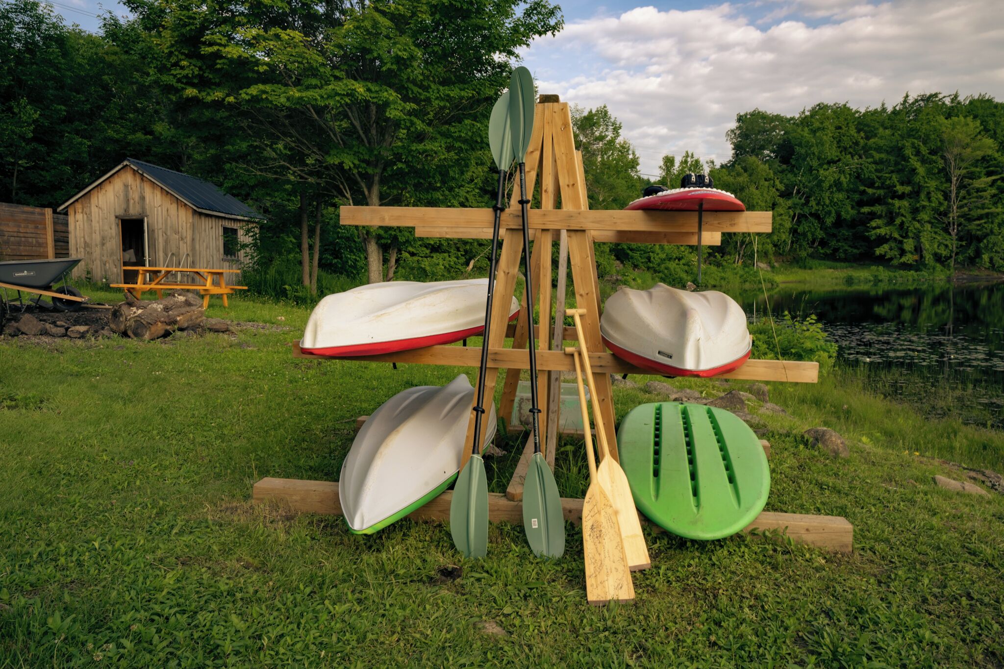 A wooden structure with kayaks and paddles, a small shed, and a grassy area near a body of water.