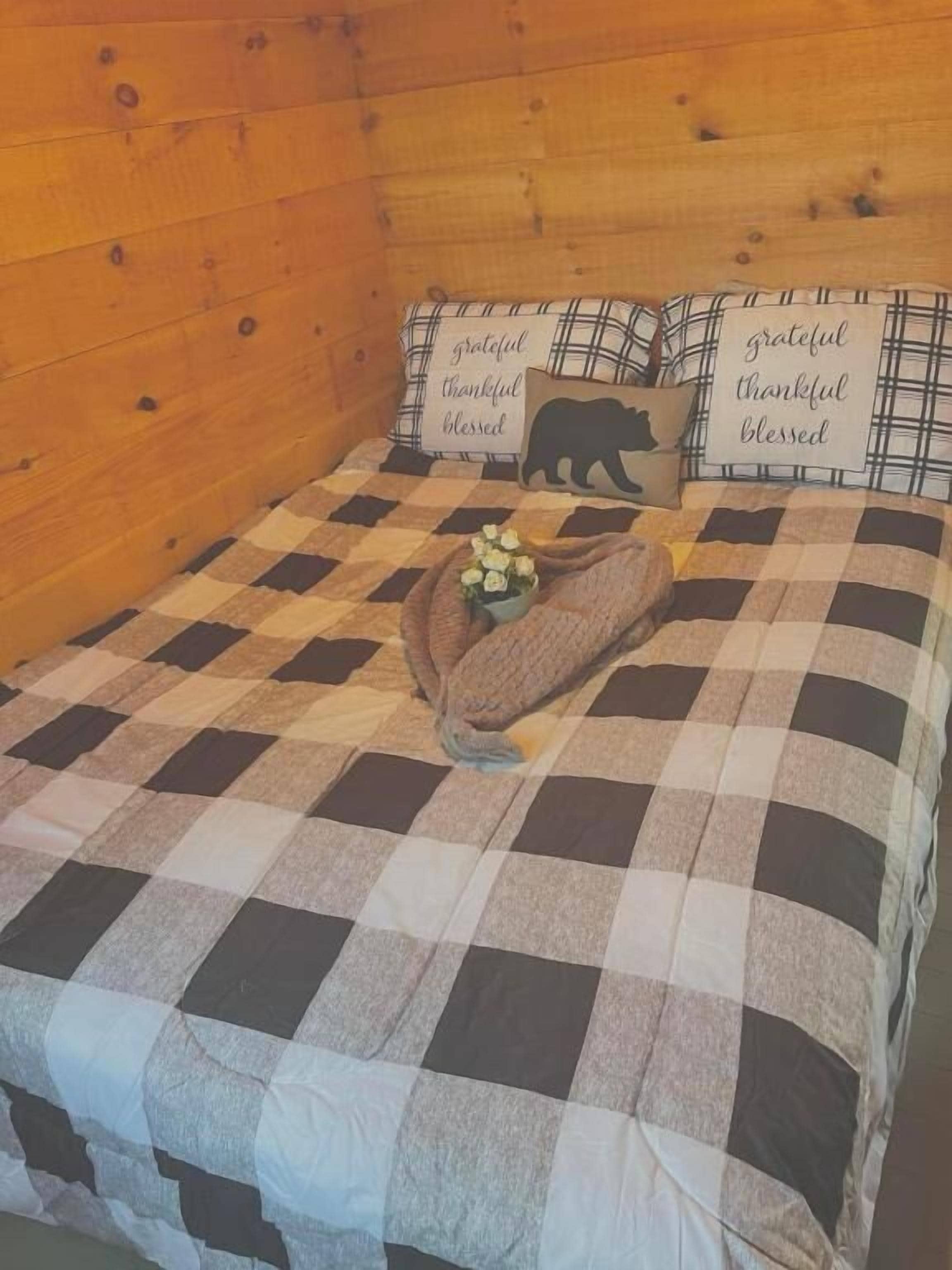 Un lit avec une couvre-lit à carreaux, deux oreillers ornés des mots « reconnaissant », « plein de gratitude » et « béni », et la silhouette d’un ours.