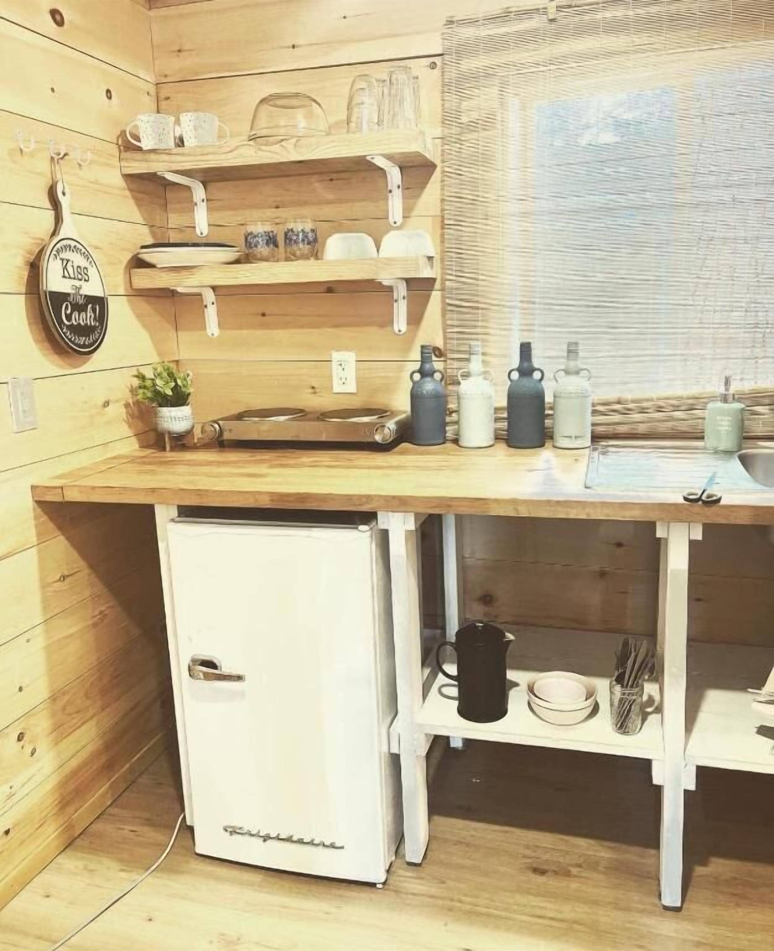 A compact kitchen with wooden walls, a white refrigerator, and shelves holding kitchenware.