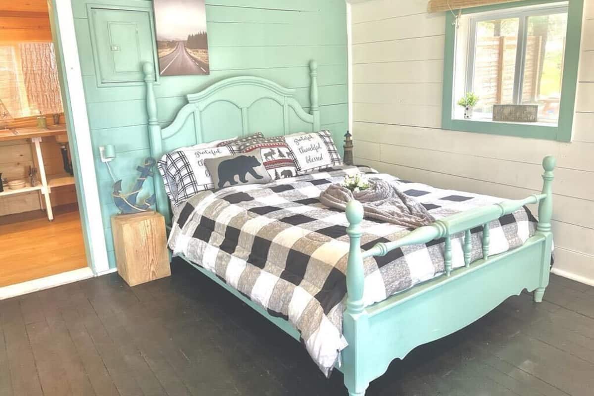 Une chambre aux murs de couleur turquoise, avec une couvre-lit à carreaux, une tête de lit décorative, un petit tabouret en bois, une étagère avec quelques objets et une fenêtre donnant sur un paysage.