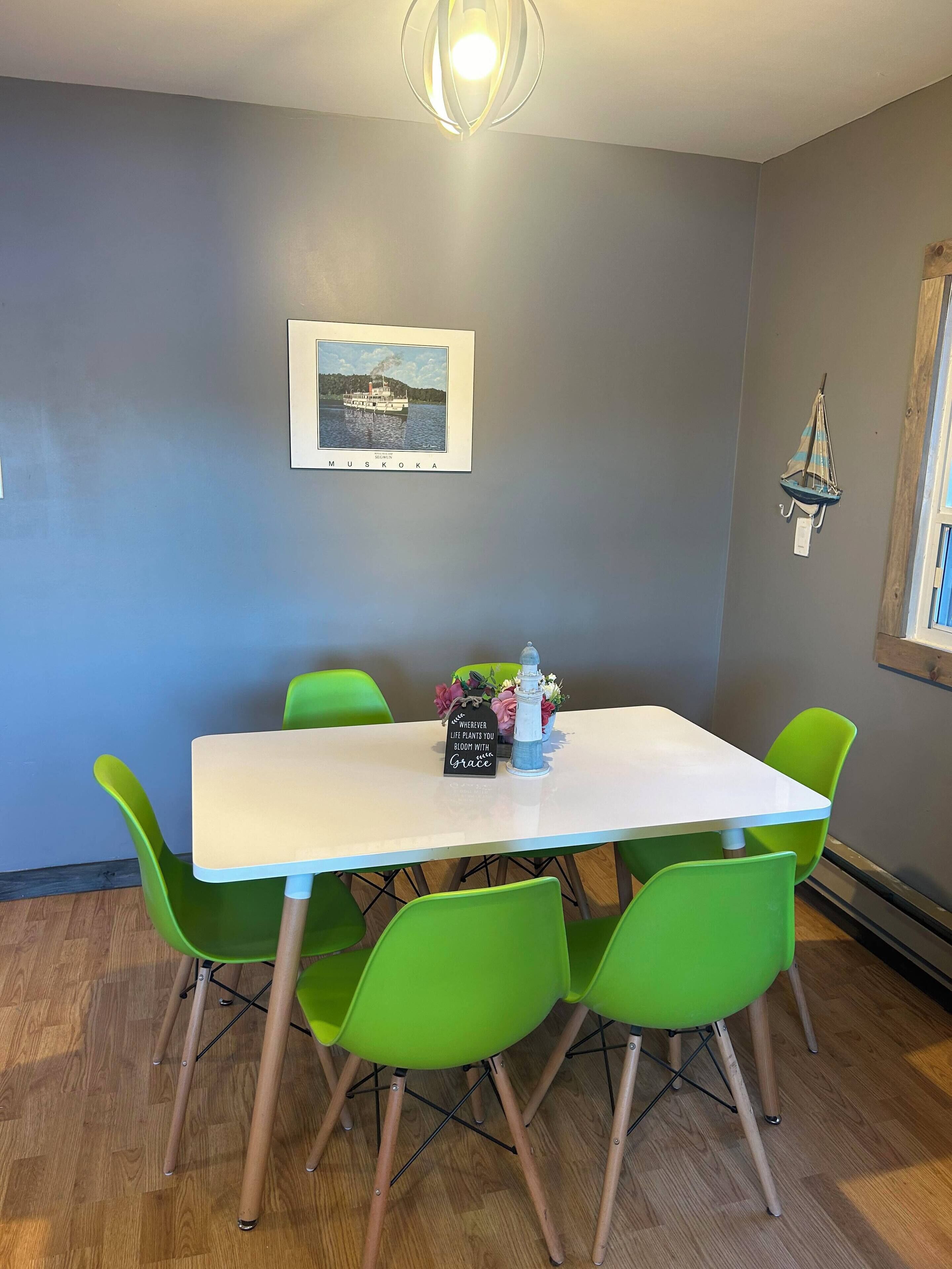 Un coin repas avec une table blanche et des chaises vertes, un tableau encadré représentant un bateau accroché au mur, et une petite décoration en forme de voilier.