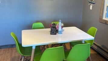 Un coin repas avec une table blanche et des chaises vertes, un tableau encadré représentant un bateau accroché au mur, et une petite décoration en forme de voilier.