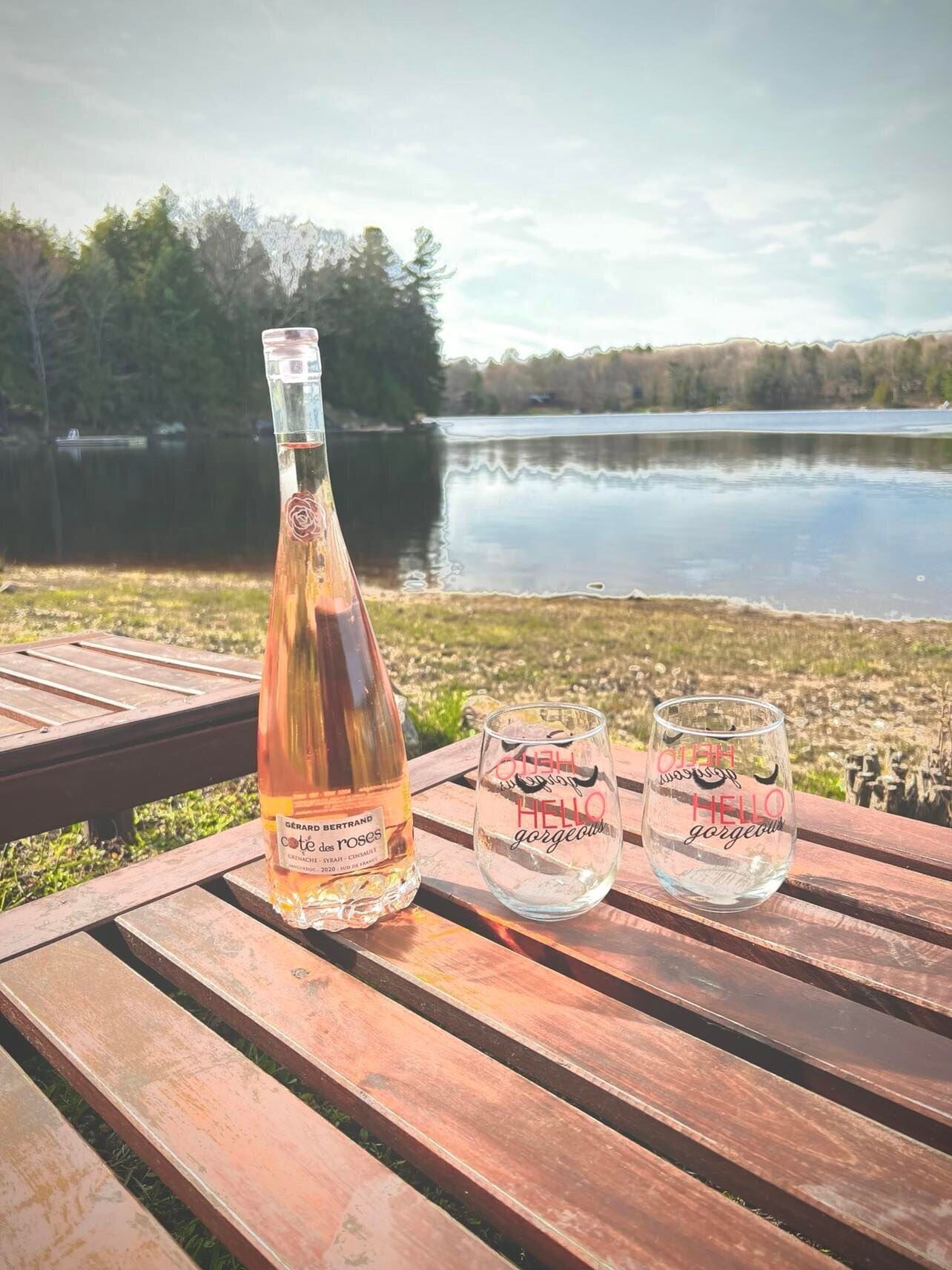A bottle of wine and two glasses on a wooden bench by a lake.
