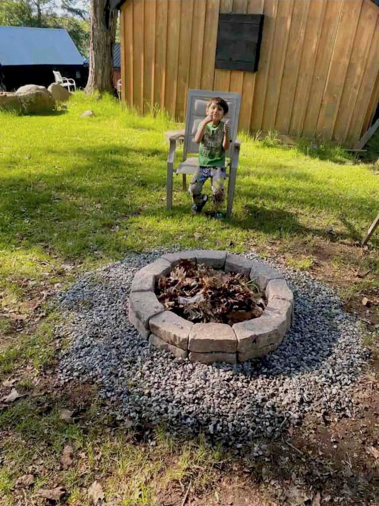 A child sitting on a chair in a backyard with a fire pit in the center.