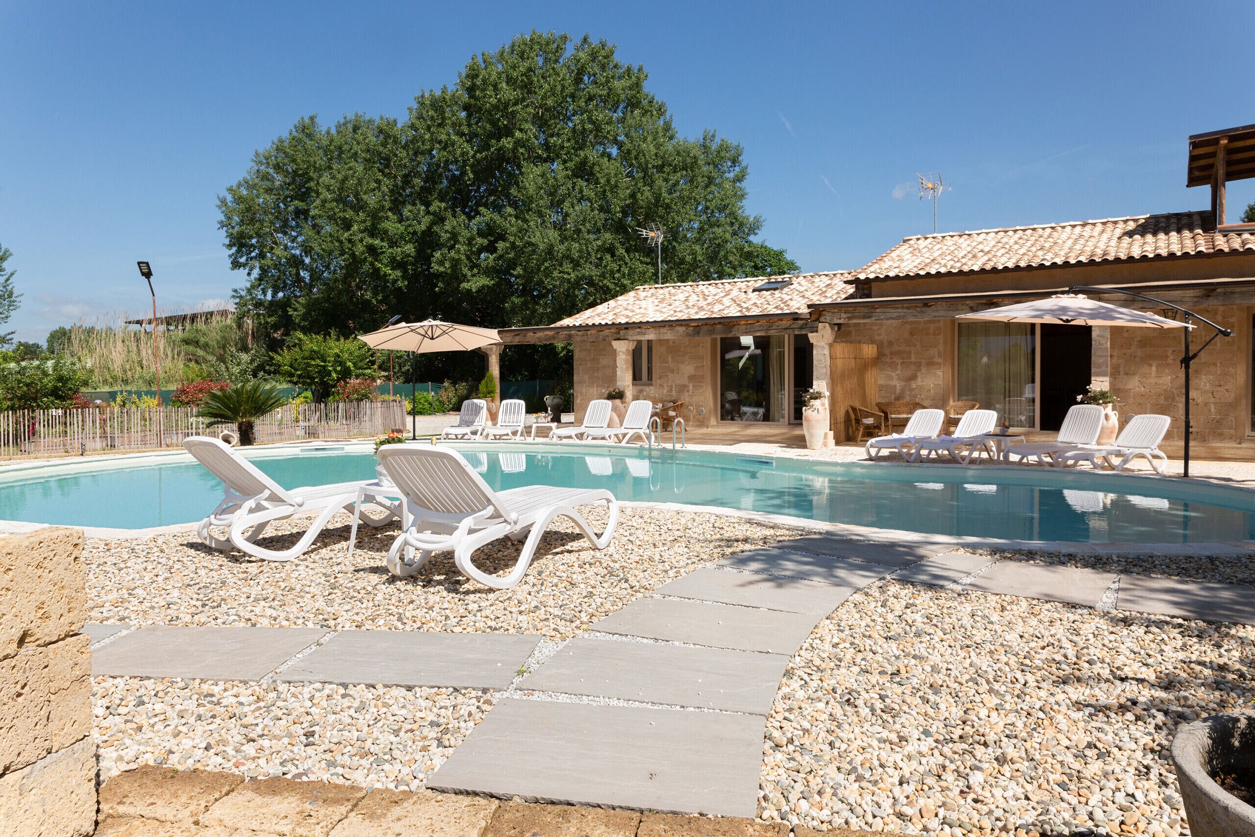 A swimming pool with lounge chairs and umbrellas, surrounded by a stone building with a tiled roof.