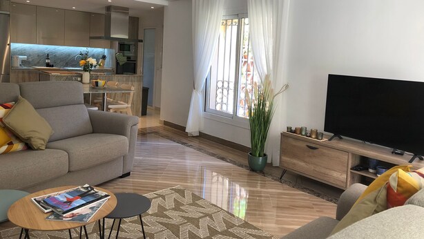 A modern living room with a sofa, a coffee table, a TV, and a kitchen area in the background.