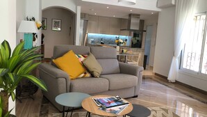 A modern living room with a grey sofa, a small round table with magazines, a potted plant, and a kitchen in the background.