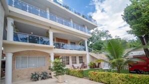 A multi-story hotel with a balcony on the upper floor, a covered outdoor seating area, and a well-maintained garden with various plants and flowers. - Fully Equipped 1BR Studio ->DT->2Mins to the Beach (sosua)