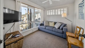 Een woonkamer met een grijze bank, een televisietoestel, een plafondventilator en uitzicht op de stad door grote ramen.