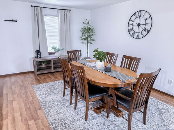 Een eetruimte met een houten tafel, zes stoelen, een klok aan de muur en een potplant.