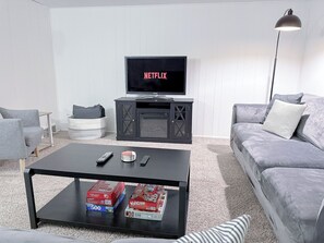 Ein Wohnzimmer mit einem Flachbildfernseher, der 'NETFLIX' zeigt, einem schwarzen Couchtisch mit Gesellschaftsspielen, einem grauen Sofa und einer Stehlampe.