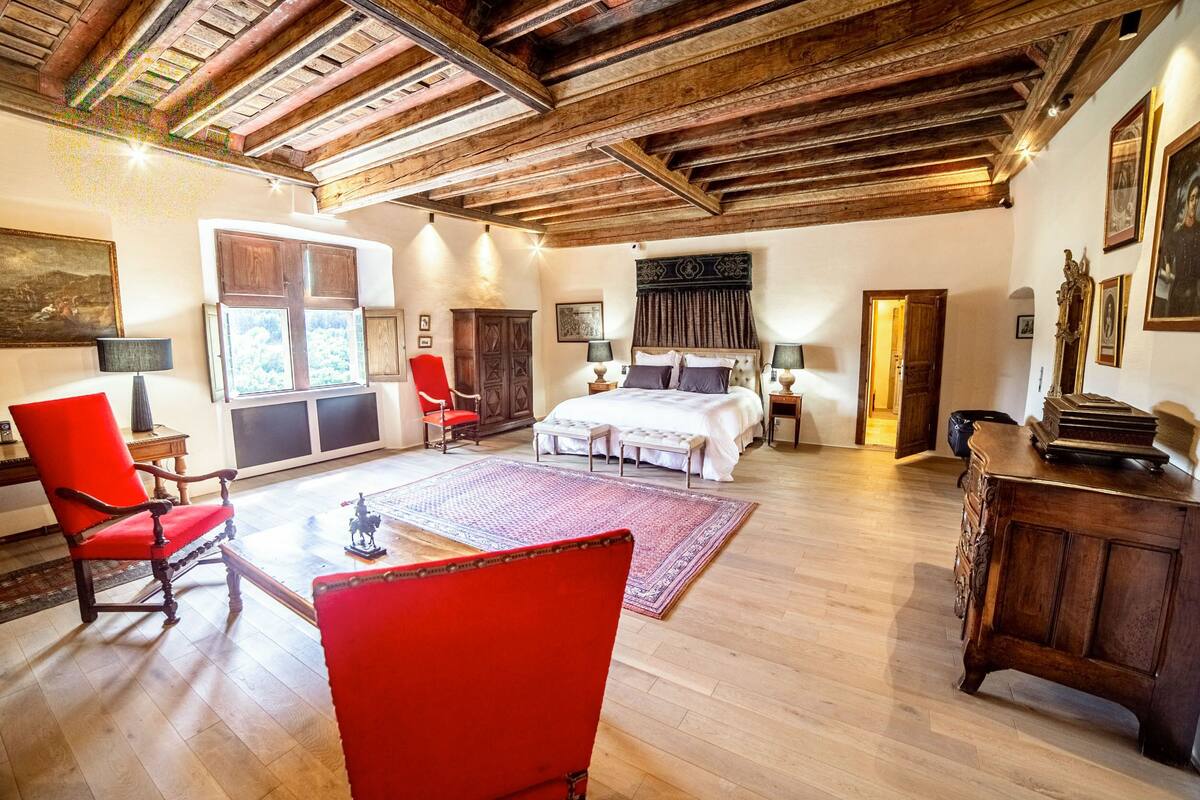 A spacious bedroom with a large bed, wooden ceiling beams, and a red chair.