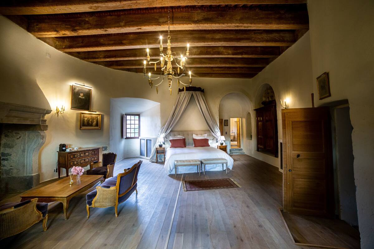 A spacious bedroom with a large bed, a chandelier, a fireplace, and a wooden ceiling.