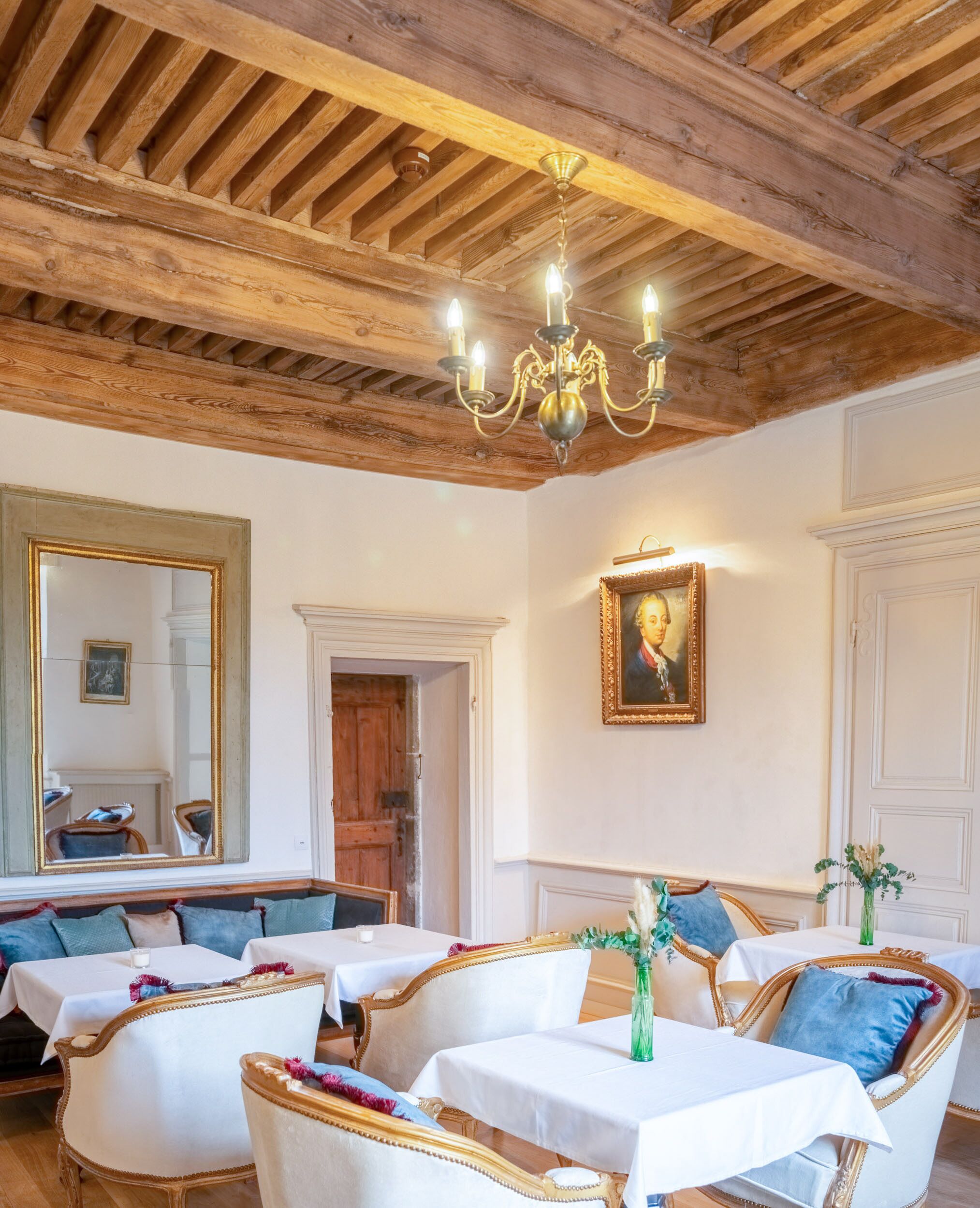 Une pièce avec des poutres apparentes, un lustre, un grand miroir, un portrait et un coin repas avec des nappes blanches et des chaises rembourrées.