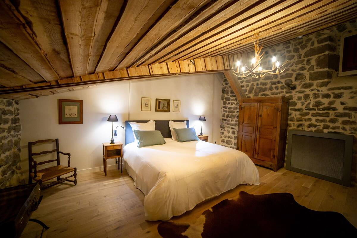 A rustic bedroom with a wooden ceiling, a stone wall, a bed with white bedding, a wooden chair, a cowhide rug, and a chandelier.