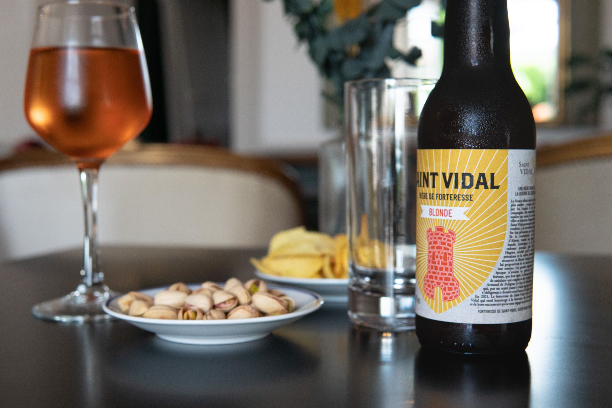 A bottle of Saint-Vidal Blonde wine, a glass of rosé wine, a plate of pistachios, and a glass of water on a table.