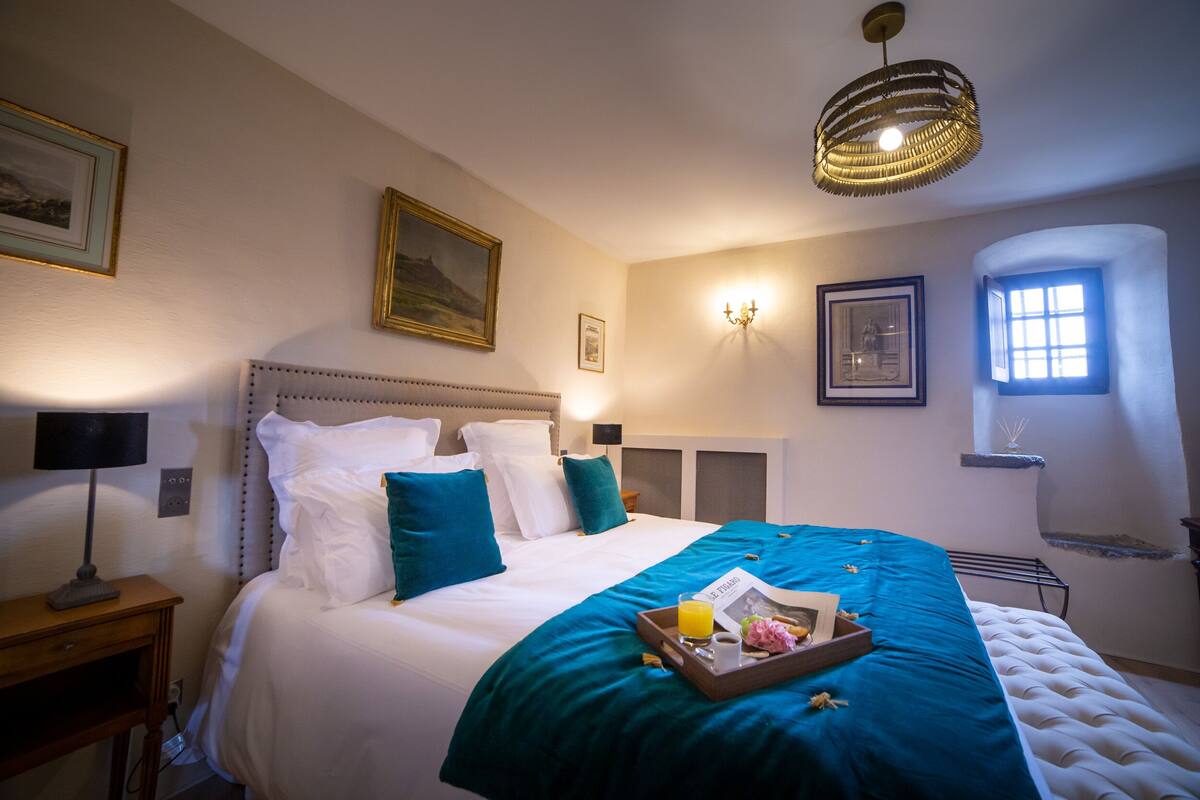 A neatly made bed with a teal bedspread, a tray with a drink and flowers, and a framed picture on the wall.