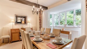 A dining area with a wooden table set for six, surrounded by chairs. A chandelier hangs from the ceiling, and there is a window with a view of greenery outside.