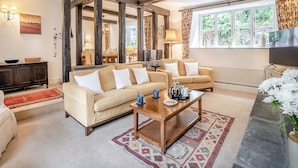 A cozy living room with a sofa set, a coffee table, and a TV. There is a window with curtains, a fireplace, and a dining area visible in the background.