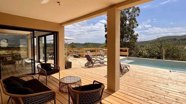 Una terraza de madera con mobiliario de mimbre, una piscina y vistas panorámicas de colinas.