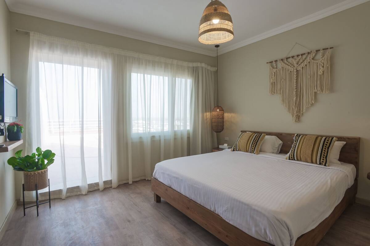 Une chambre avec un grand lit, une décoration murale en macramé suspendue, une plante en pot et un téléviseur posé sur un meuble.