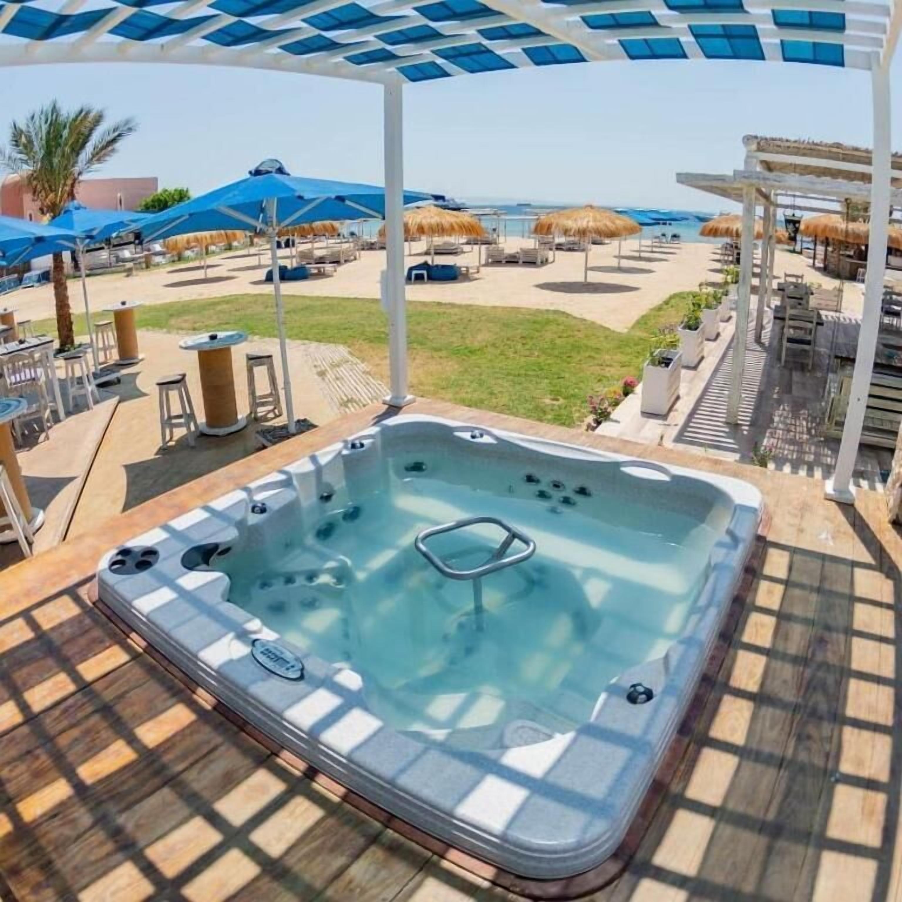 Ein Whirlpool mit Glasverkleidung, umgeben von Sitzgelegenheiten und Sonnenschirmen mit Blick auf einen Strand.