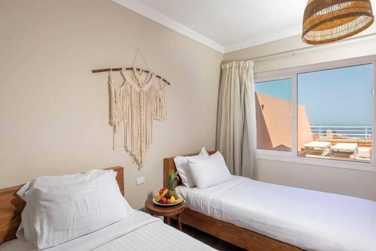 Une chambre d’hôtel avec deux lits, une décoration murale en macramé, un panier de fruits et une vue sur la mer.