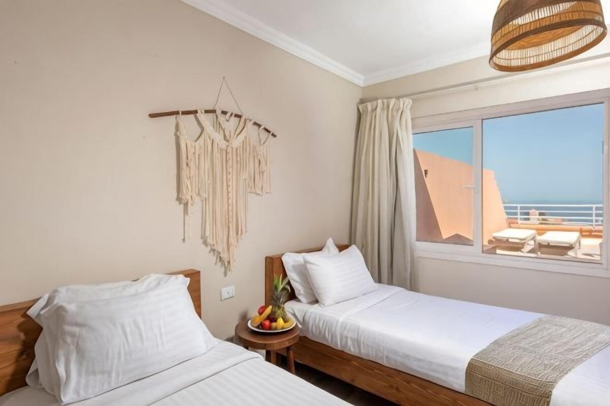 Quarto de hotel com duas camas, uma mesinha com frutas, um painel de macramé na parede e vista para o mar.