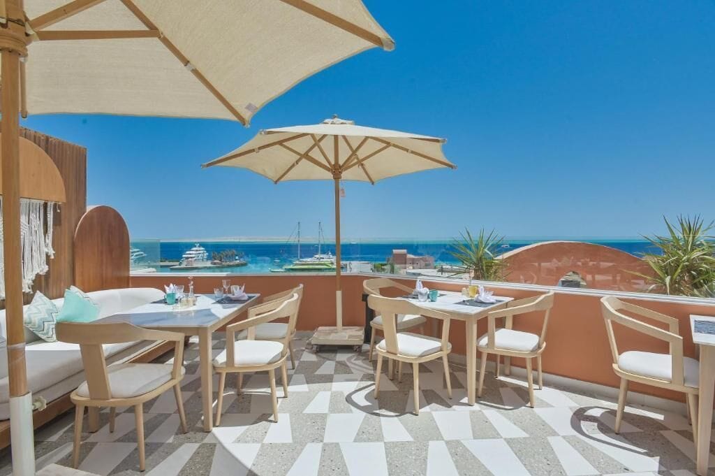 A terrace with outdoor seating and umbrellas, overlooking a coastal view with boats.