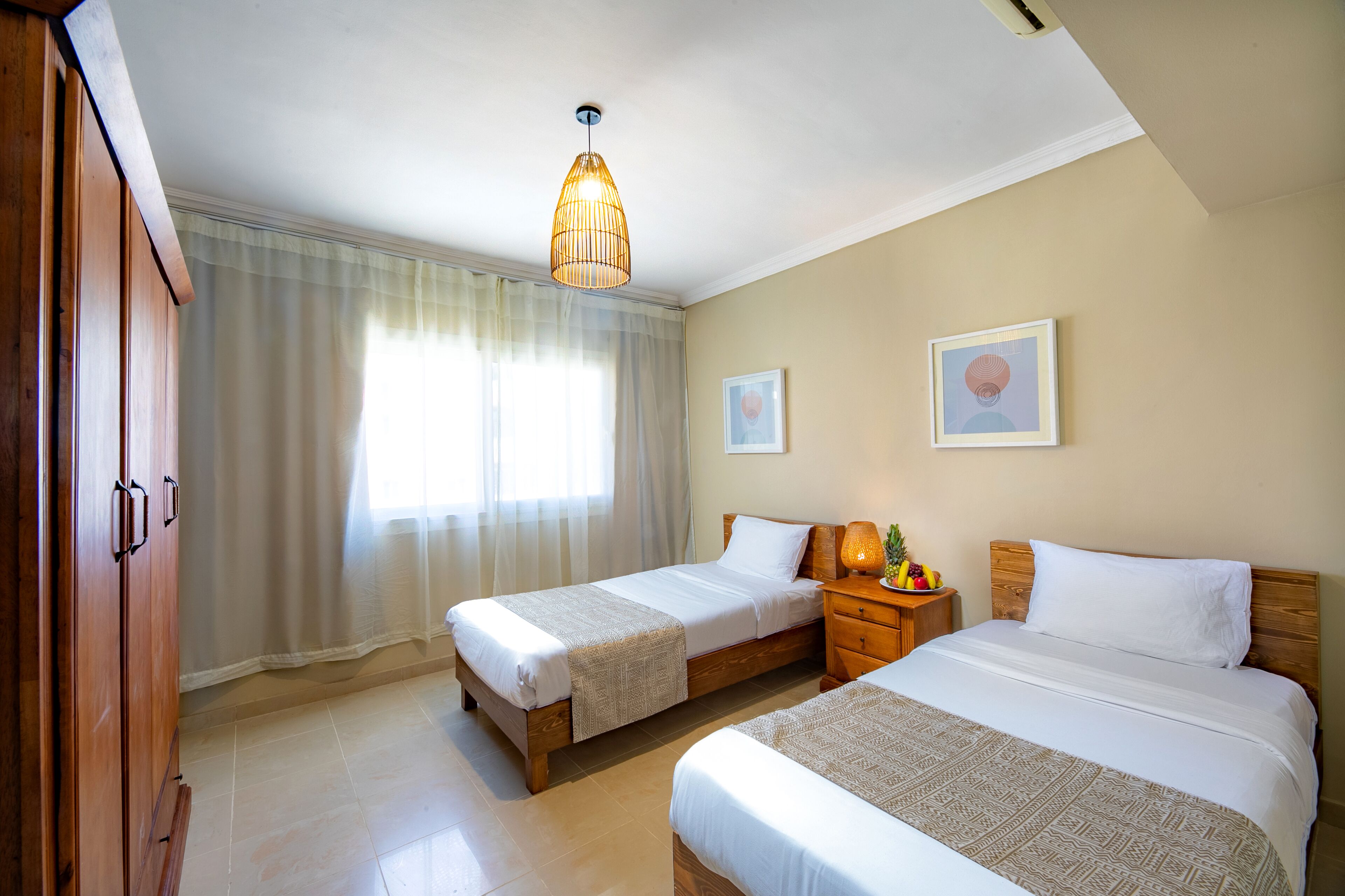 Una camera d'albergo con due letti, un armadio in legno, un comodino con una lampada e una luce a soffitto.