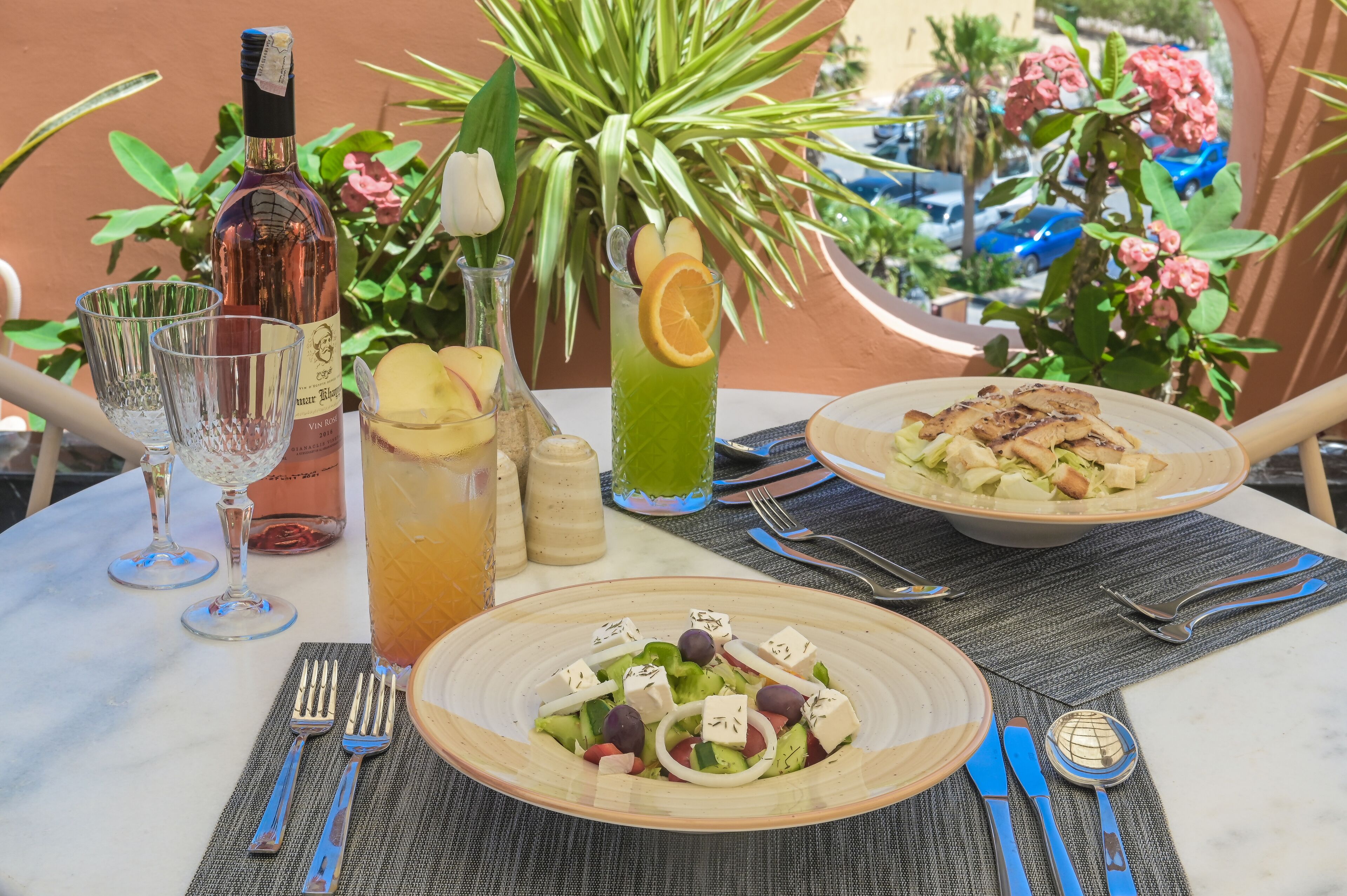 A table set with a bottle of wine, glasses, a pitcher of iced tea, a green drink with an orange slice, and plates of food including a salad and a dish with grilled vegetables.