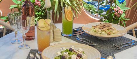 A table set with a bottle of wine, glasses, a pitcher of iced tea, a green drink with an orange slice, and plates of food including a salad and a dish with grilled vegetables.