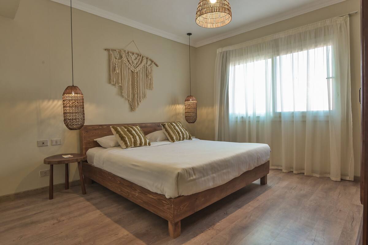 Une chambre avec un lit en bois, deux tables de chevet, une décoration murale en macramé et deux lampes suspendues.